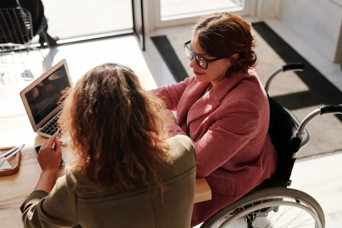 Two women working together on laptops in a bright modern workspace, one using a wheelchair