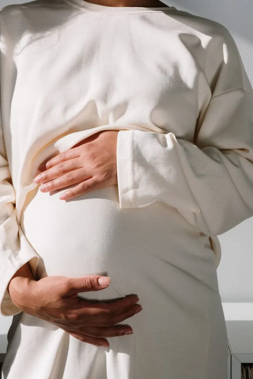 A sunlit close-up of a pregnant woman cradling her belly in a cream knit