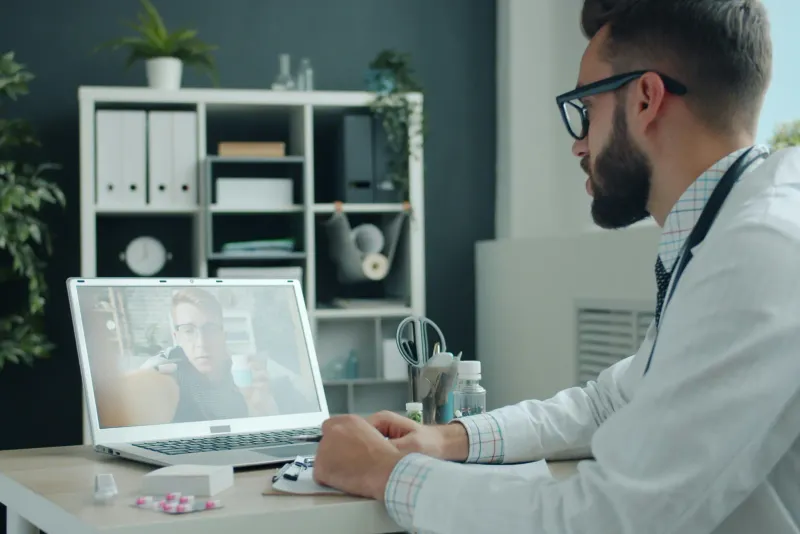A doctor at a clinic desk running a telehealth video consultation on a laptop, stethoscope beside the keyboard